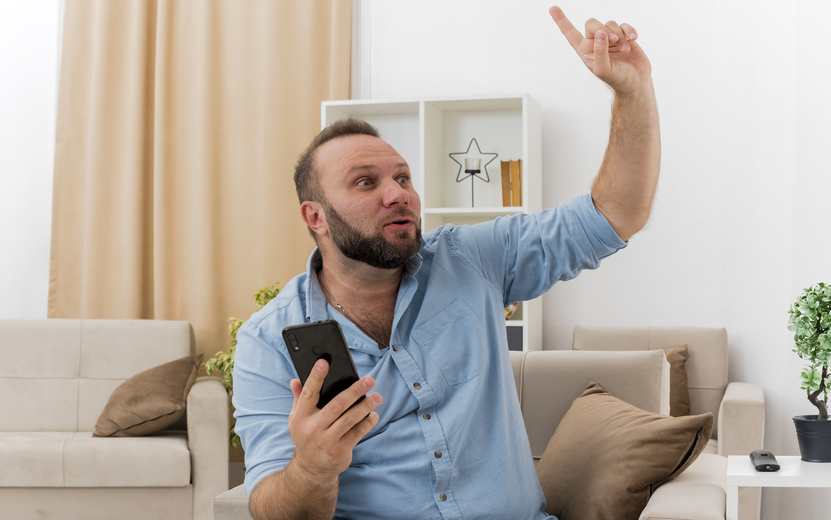 udivlennyi-vzroslyi-slavanskii-muzcina-sidit-na-kresle-derza-telefon-i-ukazyvaa-vverh-vnutri-gostinoi.jpg