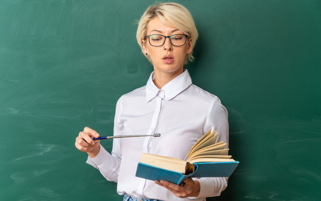 impressed-young-blonde-female-teacher-wearing-glasses-classroom-standing-front-chalkboard-holding-reading-pointing-with-pointer-stick-book-with-copy-space (1) (1).jpg