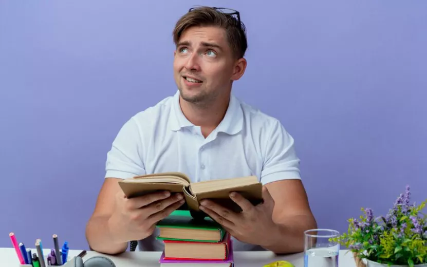 looking-side-pleased-young-handsome-male-student-sitting-desk-with-school-tools-holding-book-isolated-blue-background_141793-63049.jpg